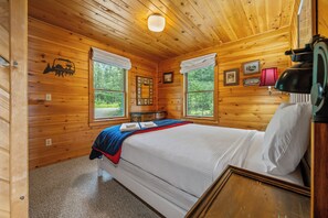 Een slaapkamer in een houten chalet, met een bed, twee ramen, een nachtkastje en een lamp.