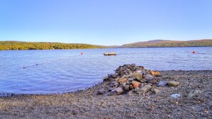 A pebbly shore with a wooden dock extending into a calm lake, surrounded by hills and clear skies. - Rangeley Manor A | Nice Cottage Close to Shared Waterfront on Rangeley Lake (Rangeley)