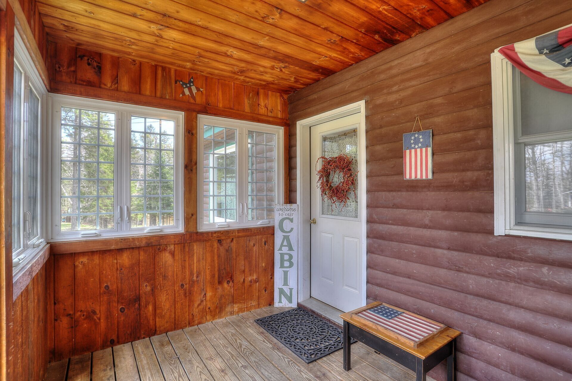 L’intérieur d’une cabane avec des murs en bois, une pancarte de bienvenue, une porte décorée d’une guirlande et un banc orné d’un drapeau américain.