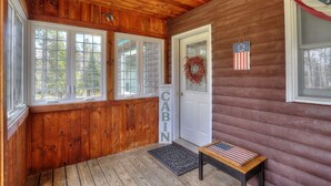 A cabin interior with wooden walls, a welcome sign, a door with a wreath, and a bench with an American flag. - Rangeley Manor A | Nice Cottage Close to Shared Waterfront on Rangeley Lake (Rangeley)