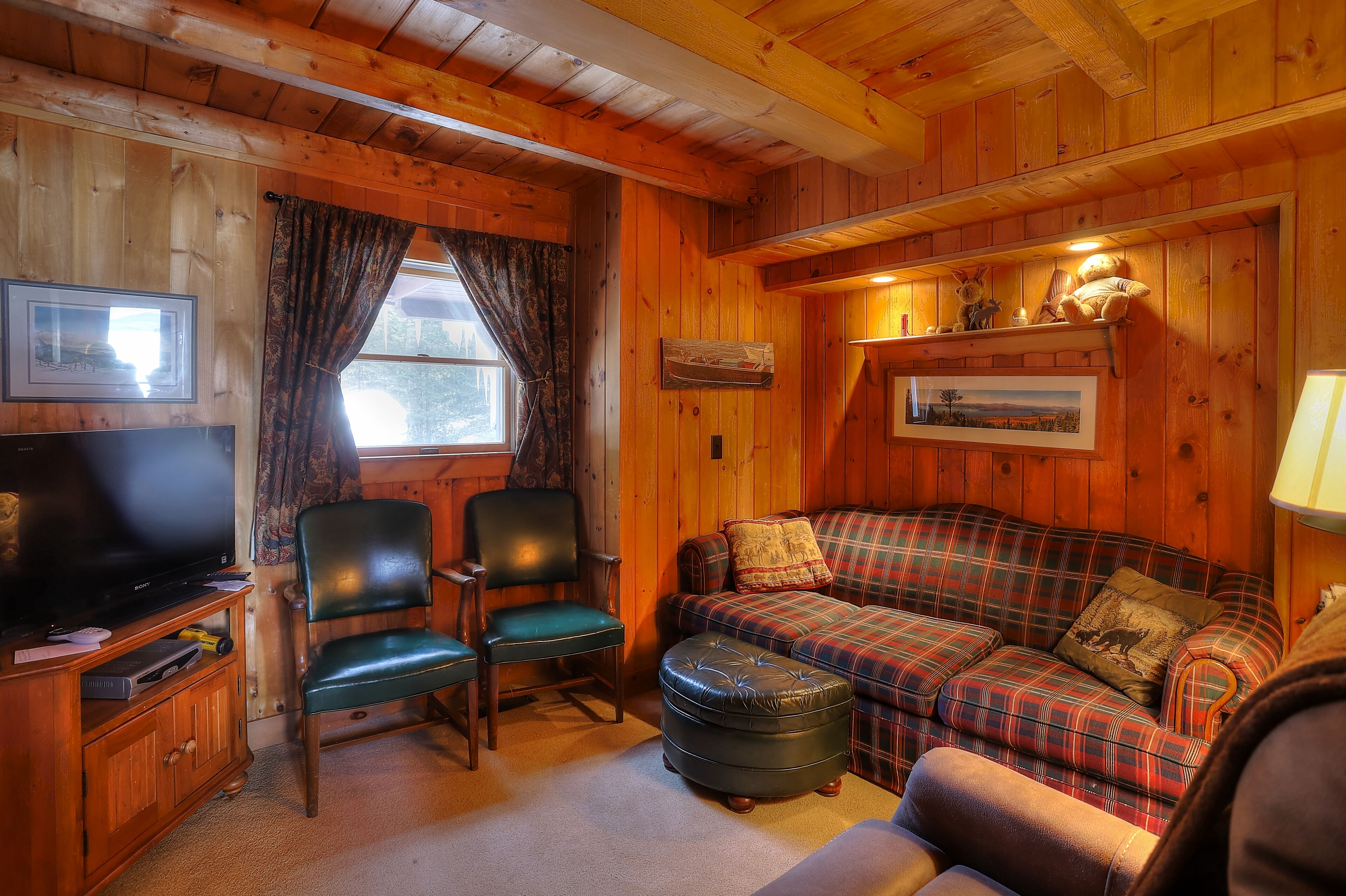 A cozy cabin interior with a plaid sofa, green chairs, a television, and wooden walls.