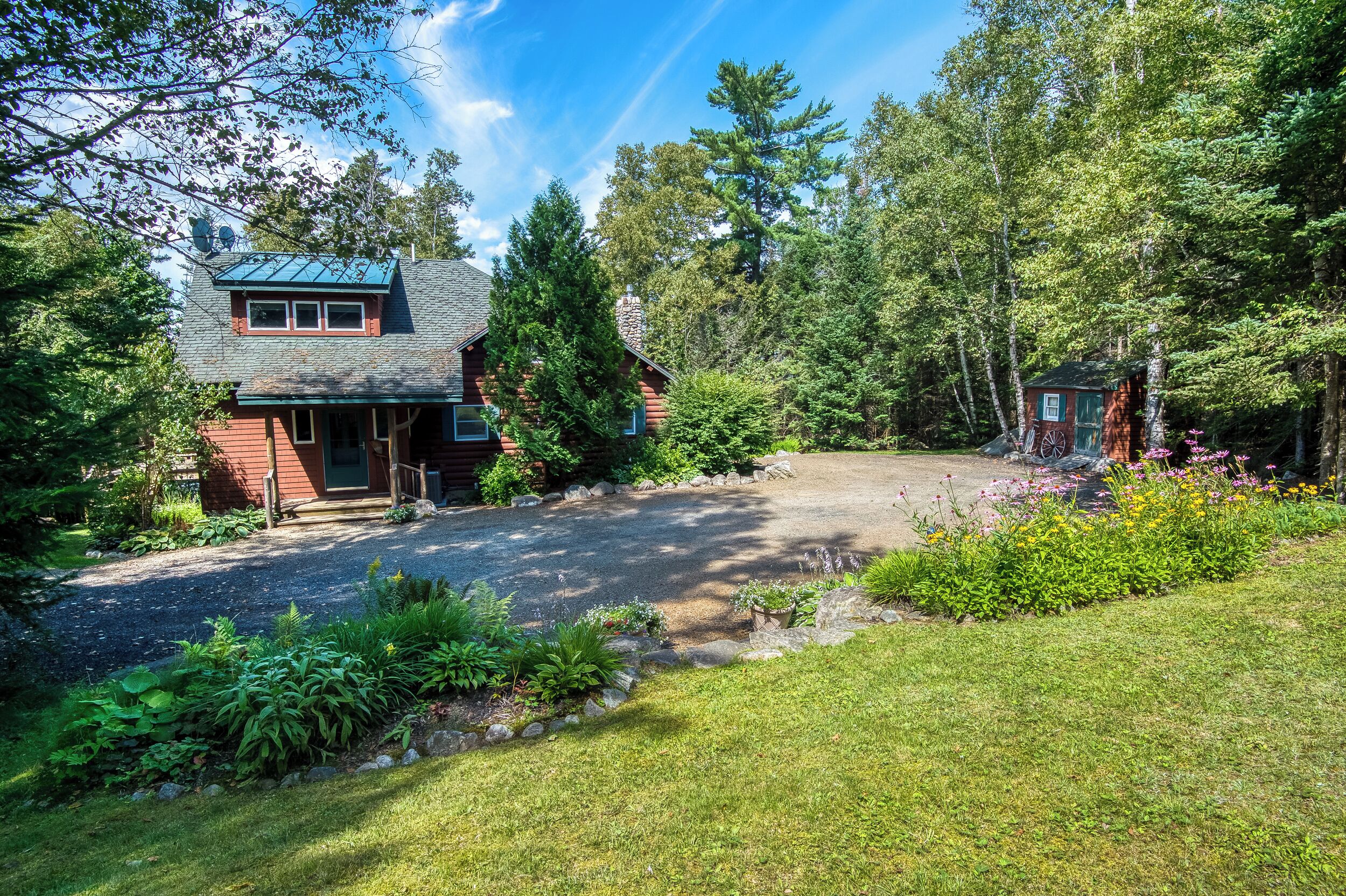 A log cabin with a stone foundation, surrounded by a well-maintained garden and dense forest.