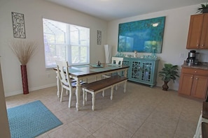 A dining area with a table and chairs, a window with blinds, a painting on the wall, and a kitchen area with cabinets and appliances.