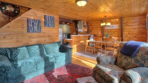 Interior d'una cabana acollidora, amb sostre de fusta, zona de menjador amb taula i cadires, un sofà i una petita cuina.