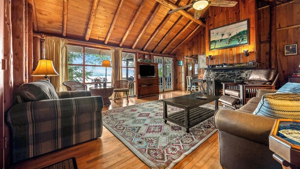 A cozy log cabin interior with a fireplace, wooden ceiling, and a large window overlooking a scenic view. - 🐾Kadel - Maine log home on Mooselookmeguntic Lake filled with rustic charm, dog friendly (Oquossoc)