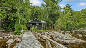 A wooden bridge leading to a cabin surrounded by dense forest and a clear stream. - 🐾Kadel - Maine log home on Mooselookmeguntic Lake filled with rustic charm, dog friendly (Oquossoc)