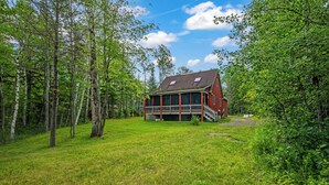 Une grande maison en bois à deux étages, dotée d’un porche, entourée d’un paysage verdoyant et luxuriant et de grands arbres.