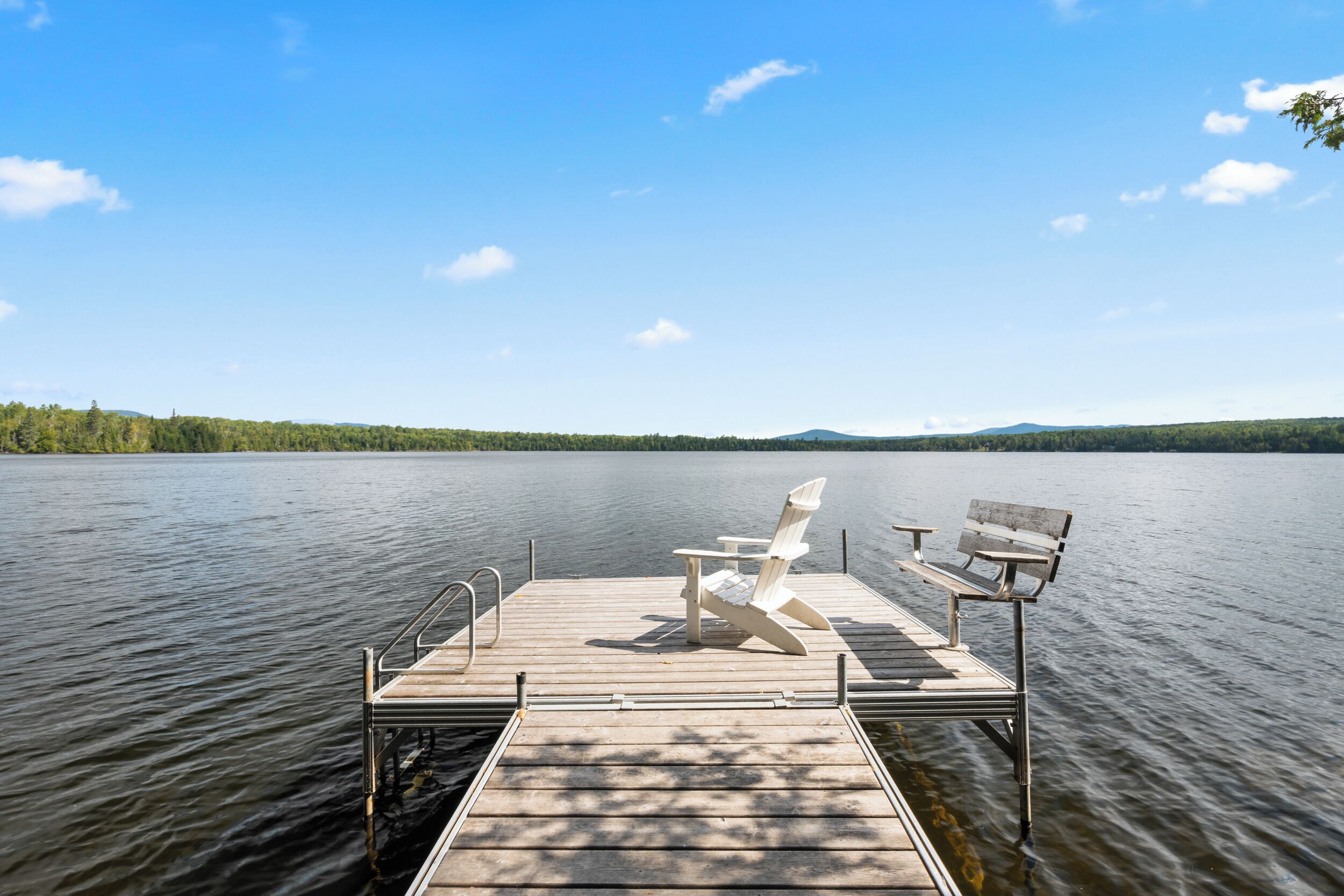 A wooden dock with two Adirondack chairs and a bench, extending over calm water with a forested shoreline in the background.