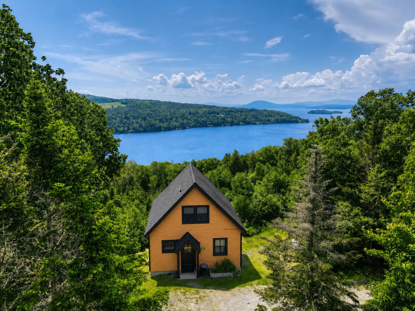 Eine Holzhütte mit Satteldach, umgeben von dichtem Wald, mit Blick auf ein großes Gewässer und ferne Hügel.