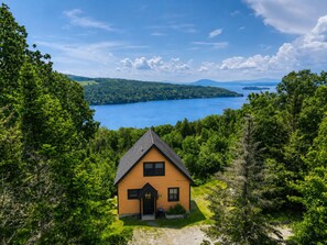 A wooden cabin with a gabled roof, surrounded by dense forest, overlooking a large body of water and distant hills.