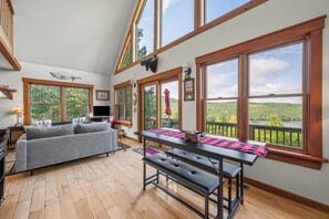 A spacious room with a wooden floor, a grey sofa, a dining table with benches, and large windows offering a view of a forested landscape.