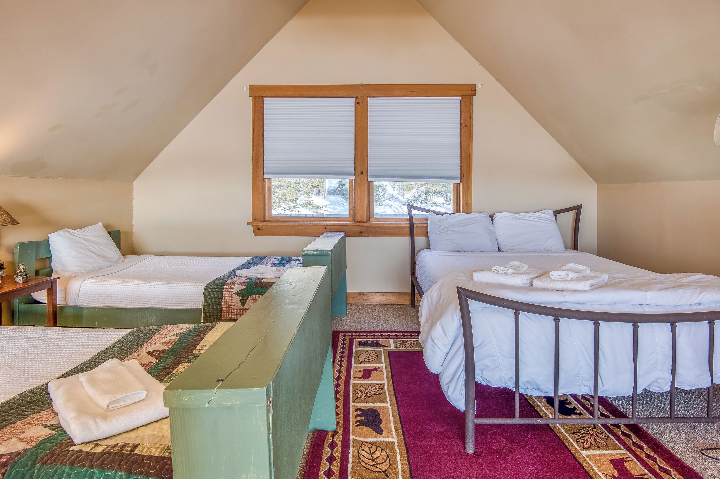 A bedroom with two beds, a window with blinds, a wooden ceiling, and a red and green patterned rug.