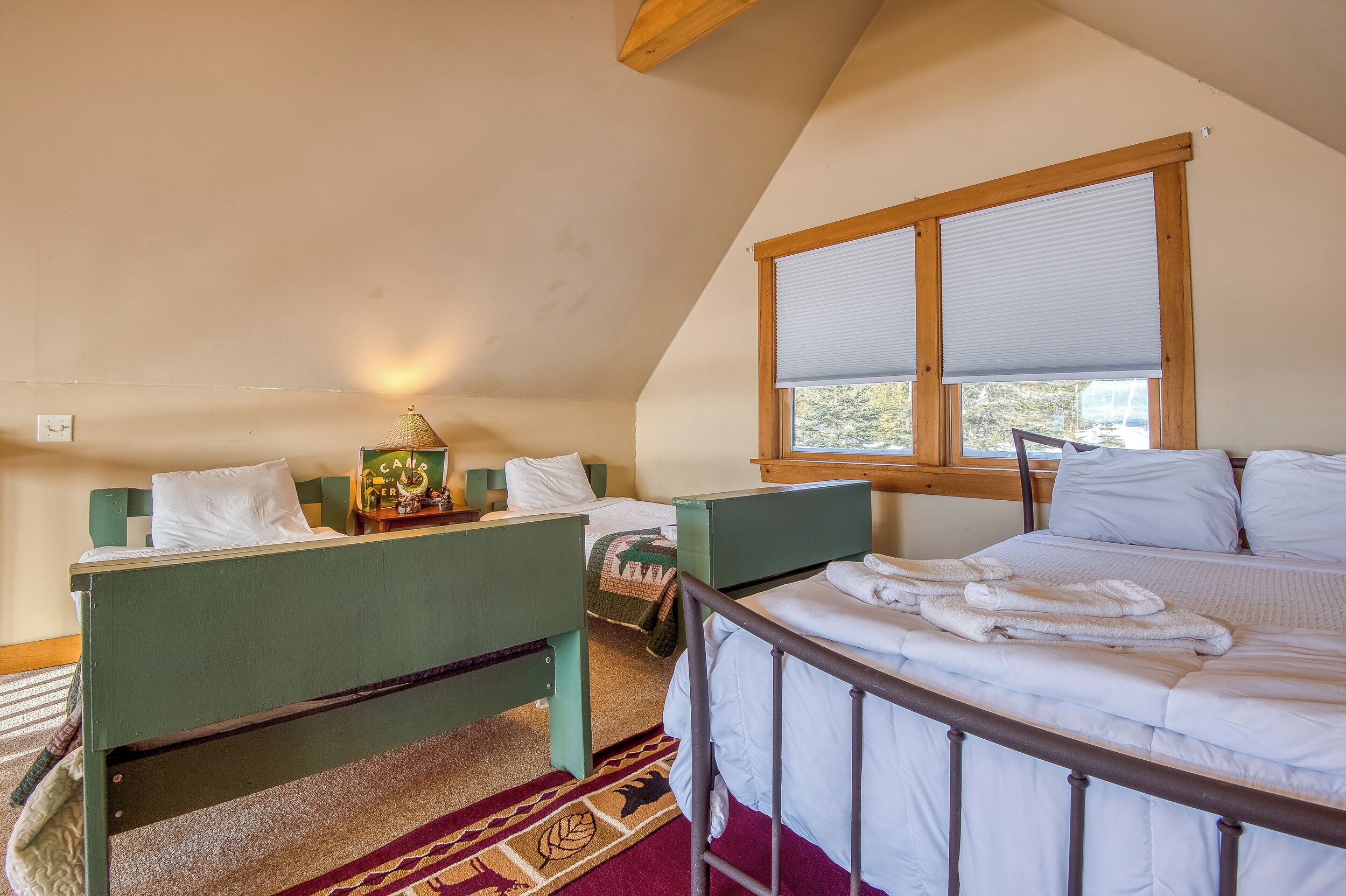 A bedroom with two single beds, a window with blinds, a small table with a lamp, and a carpeted floor.