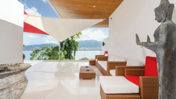 Eine moderne Lounge im Freien mit Rattanmöbeln, einer großen Steinskulptur und Blick auf Berge und Wasser.