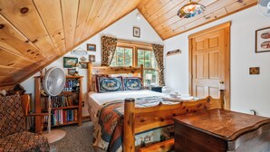 A cozy room with a wooden ceiling, a bed with a quilt, a wooden chair, a table, a bookshelf, and a window with curtains. - Alpine Loon Lodge - Just a mile from downtown Rangeley with lake views! (Rangeley)