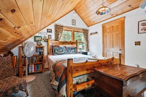 Una camera accogliente con soffitto in legno, un letto con copriletto, una sedia di legno, un tavolo, una libreria e una finestra con tende.