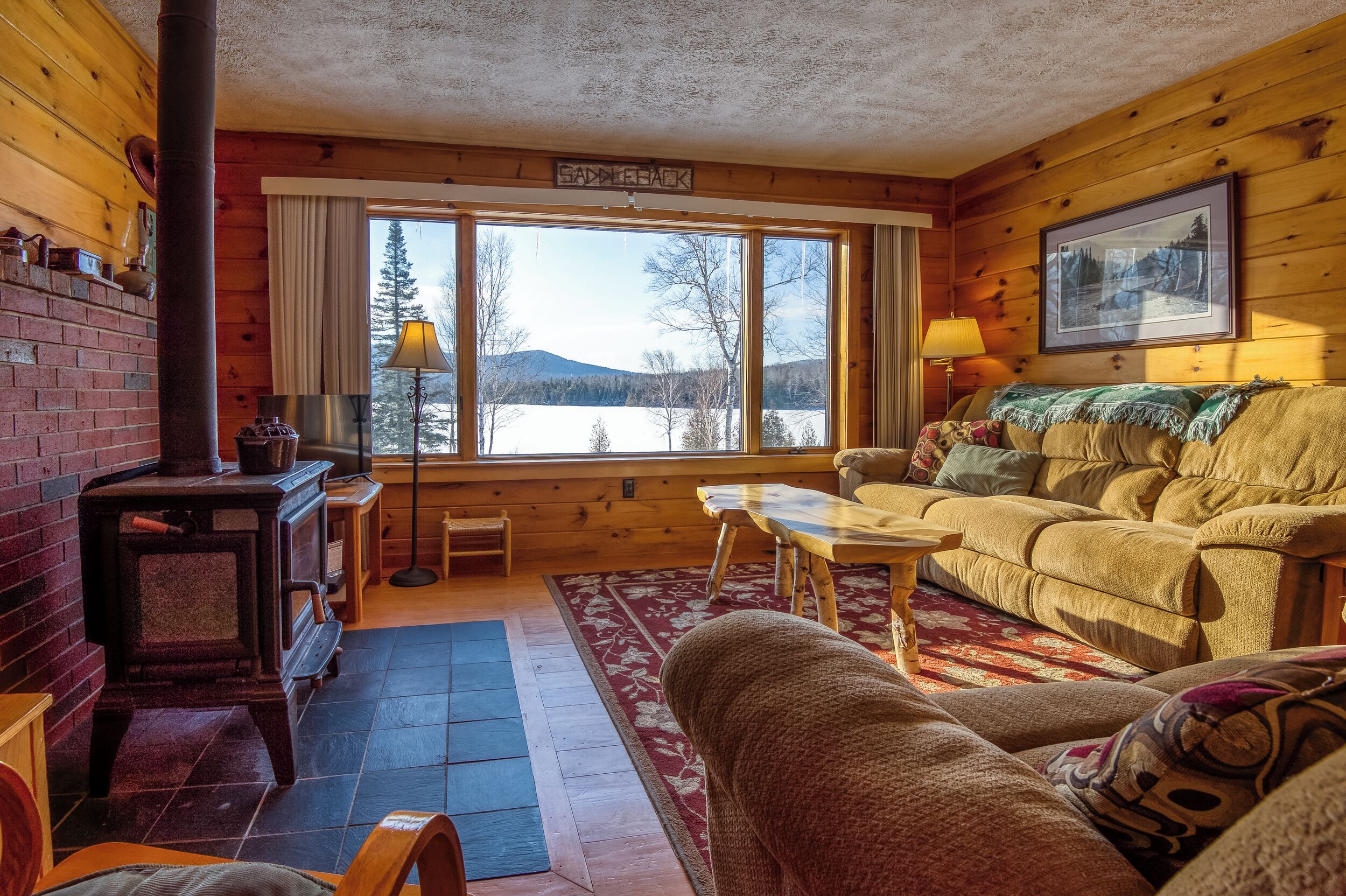 Interior acogedor de una cabaña con chimenea, un sofá, una mesa y vistas a un lago y montañas a través de amplias ventanas.