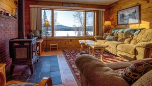 L’intérieur chaleureux d’un chalet avec une cheminée, un canapé, une table et une vue sur un lac et des montagnes, le tout visible à travers de grandes fenêtres.