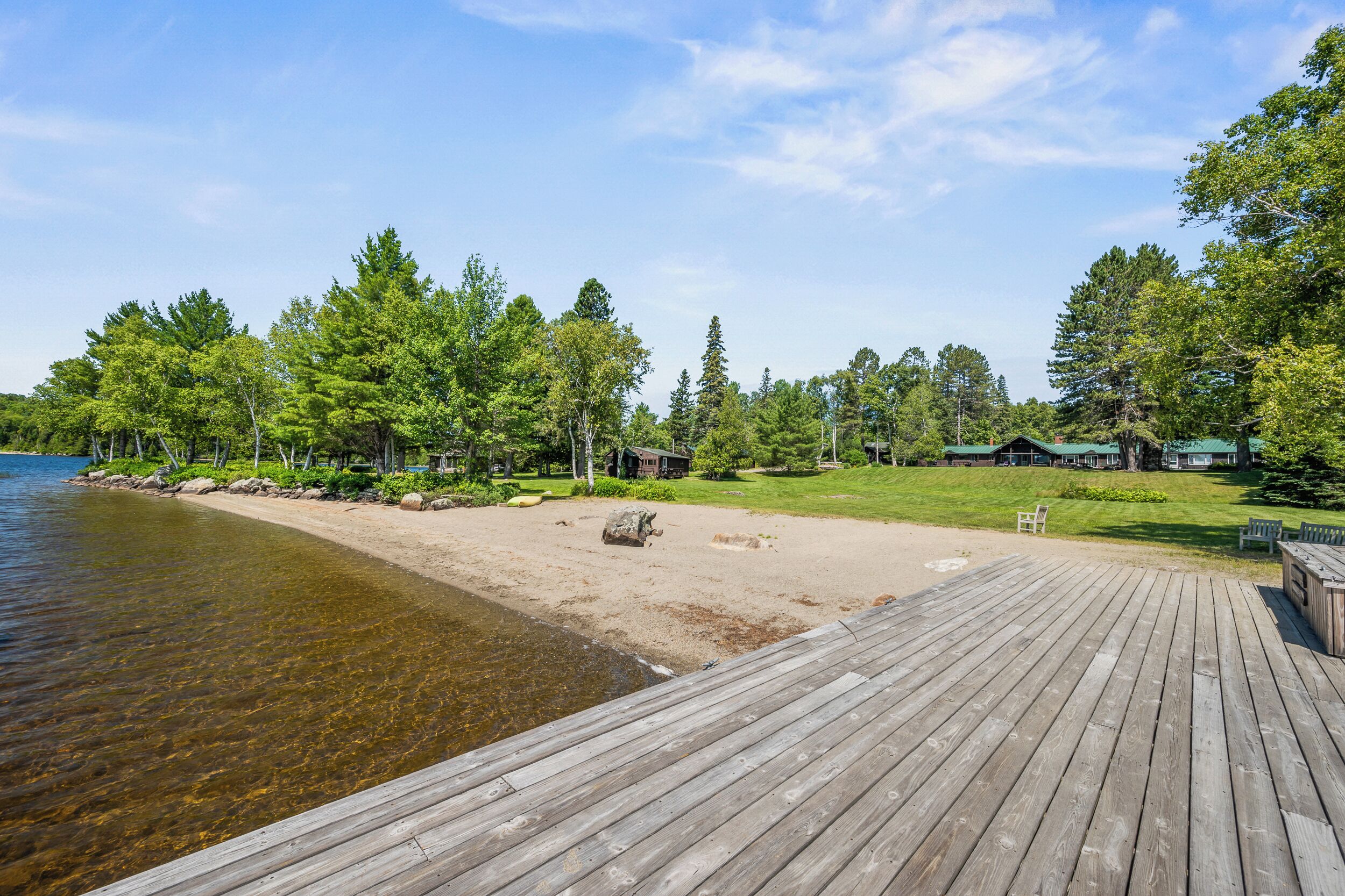 Een houten terras leidt naar een zandstrand met helder water, omgeven door bomen en een chalet.