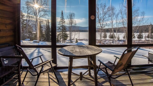 Une terrasse en bois avec une table et des chaises, une grande fenêtre donnant sur un paysage enneigé et des arbres.