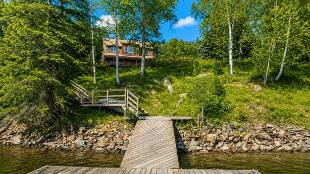 Une jetée en bois menant à une maison dotée d’un balcon, entourée d’arbres et bordée par un plan d’eau.
