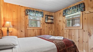 Une pièce aux murs lambrissés, avec un lit, une fenêtre avec des rideaux, une lampe fixée au mur et une petite chaise en bois.