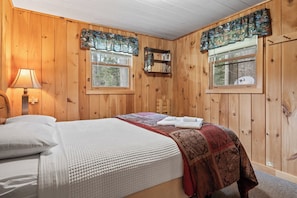 Una stanza con rivestimento in legno, un letto, una finestra con tende, una lampada a parete e una piccola sedia di legno.