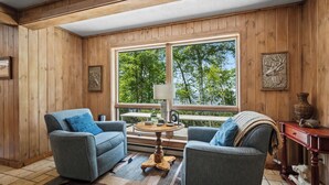 A wooden-paneled room with two blue armchairs, a small wooden table, and a large window overlooking trees.