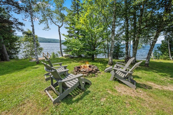 A lakeside setting with Adirondack chairs around a campfire, surrounded by trees and a view of the lake.