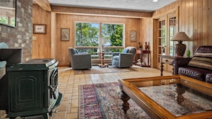 A cozy living room with a fireplace, two armchairs, a coffee table, and a large window offering a view of trees.