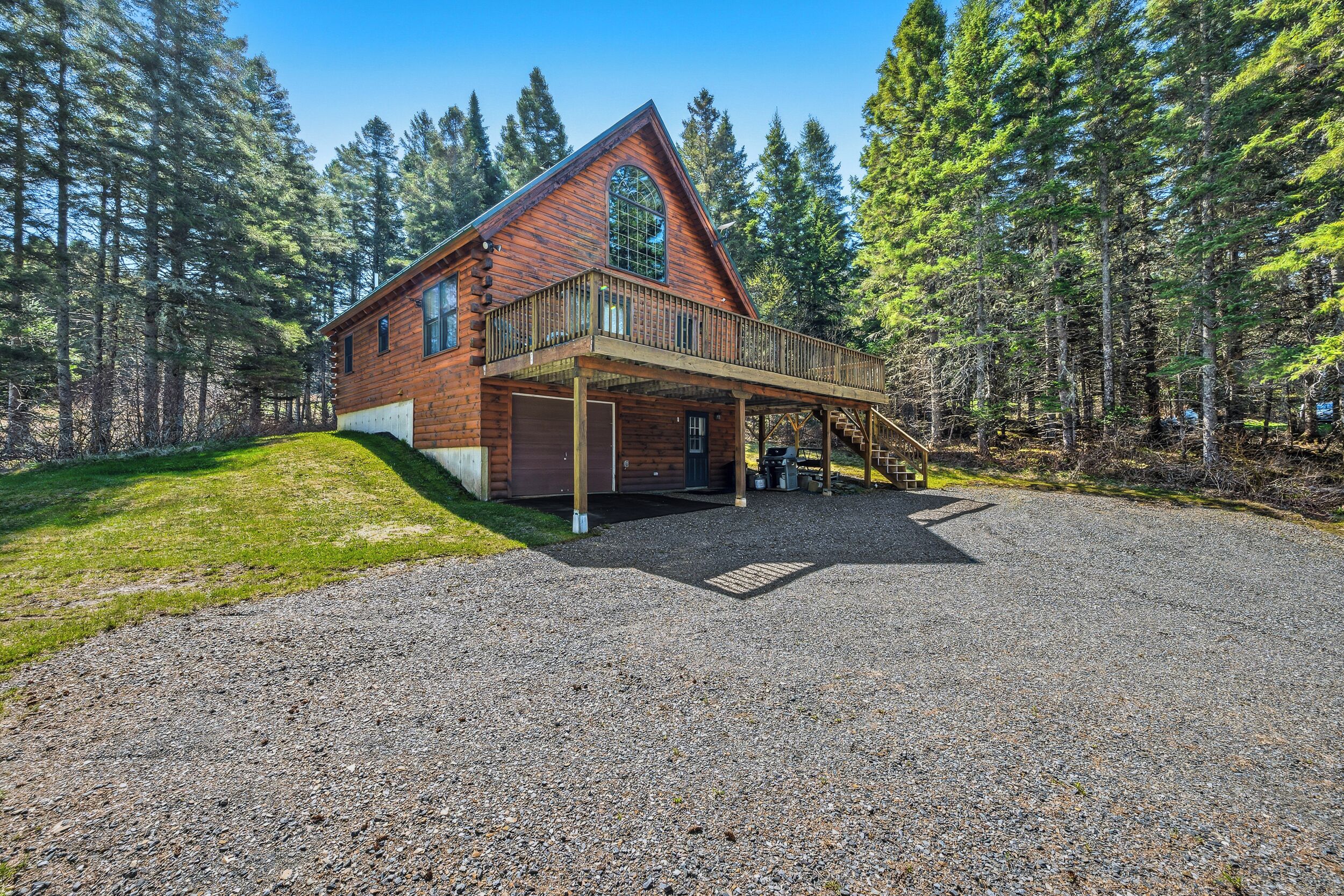 A log cabin with a covered porch, surrounded by a gravel driveway and dense forest.