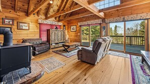 A wooden cabin interior with a fireplace, sofa, armchair, and large windows. - 🐾33 Loop Rd Log Cabin w/ Shared Rangeley Lake Access, Snowmobile/ATV Access, 15 min to Saddleback (Rangeley)