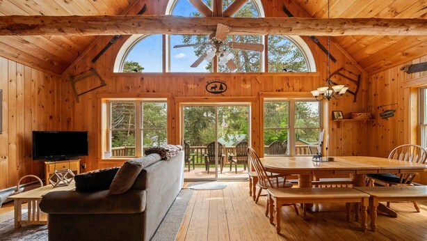 A wooden cabin interior with a sofa, dining table, chairs, a TV, and large windows. - Russell Cove 18 | Pet-Friendly Log Home | Lake Access | Sleeps 10 (Rangeley)