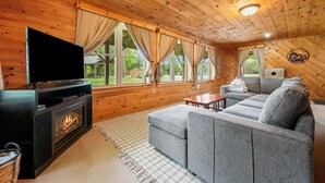 A cozy log cabin interior with a fireplace, a flat-screen TV, a gray sofa, and large windows offering a view of the outdoors. - Russell Cove 18 | Pet-Friendly Log Home | Lake Access | Sleeps 10 (Rangeley)