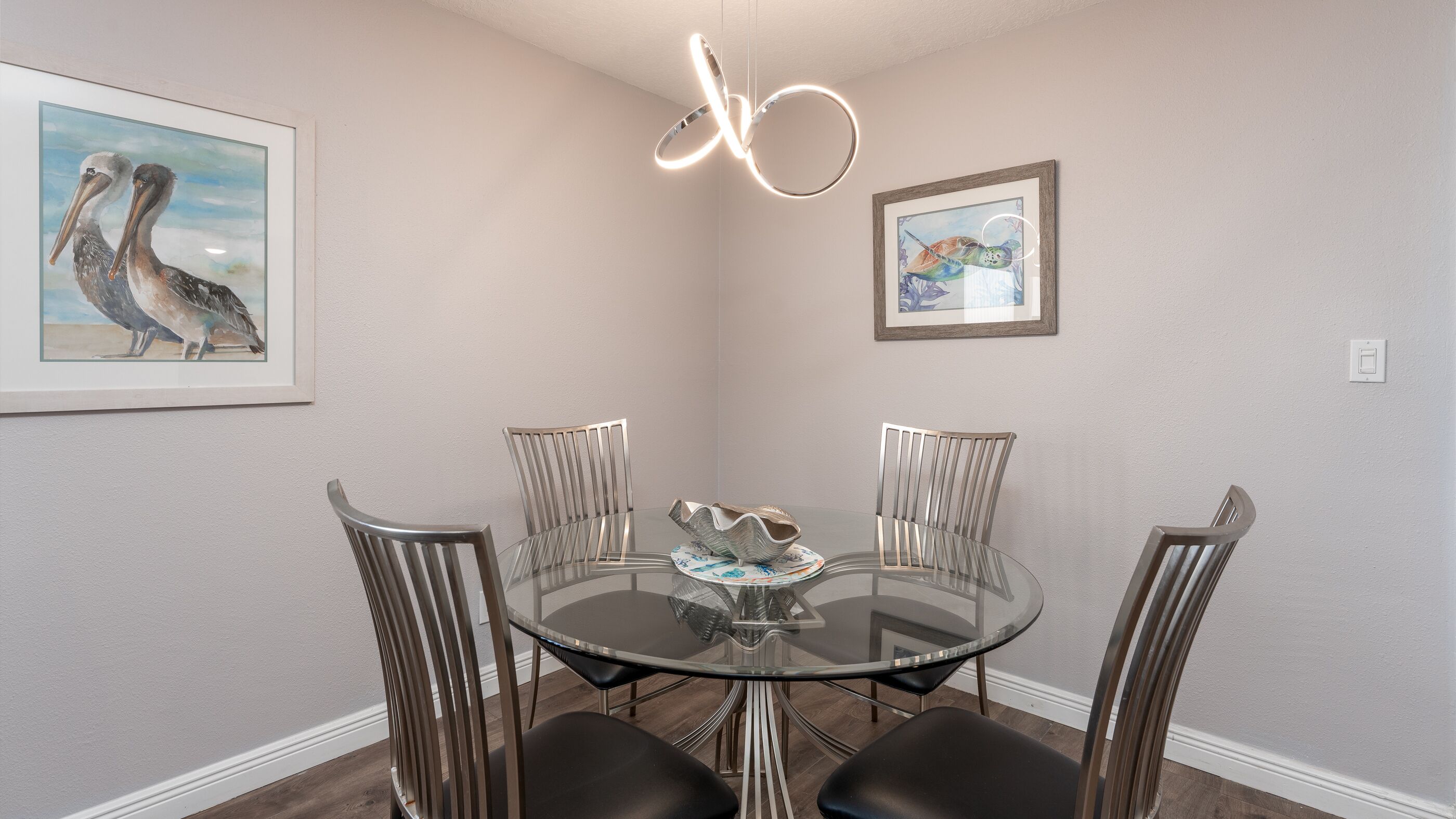 A dining area with a round glass table, four chairs, and two framed artworks on the walls.
