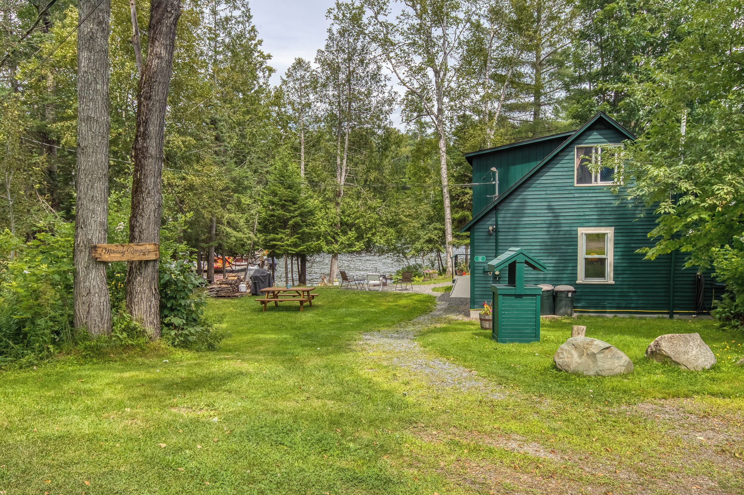 A green cabin with a picnic table and a small shed, surrounded by trees and a gravel path.
