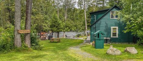 A green cabin with a picnic table and a small shed, surrounded by trees and a gravel path.