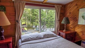 Habitación tipo cabaña de madera con cama, mesita de noche, lámpara y ventana con vistas a un bosque.