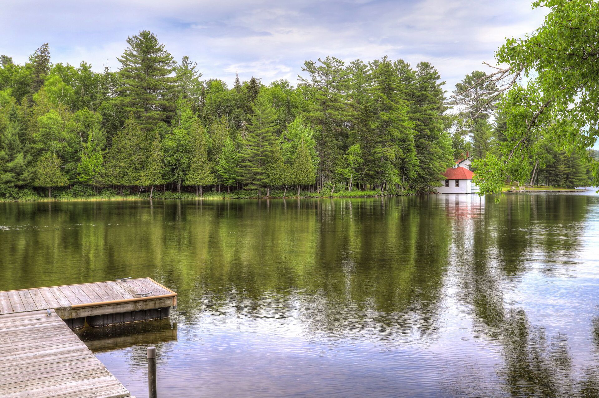 A calm lake with a wooden dock extending into the water. In the background, there is a small house with a red roof near the water's edge, surrounded by dense green trees.