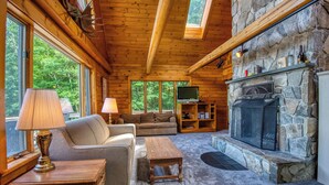 L’intérieur chaleureux d’un chalet en rondins, avec une cheminée en pierre, des meubles en bois et de grandes fenêtres donnant sur la forêt.