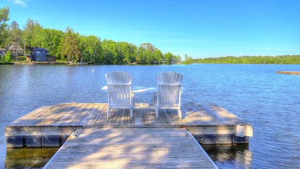 A wooden dock with two Adirondack chairs facing a calm lake, surrounded by trees and a house in the background. - Village Walk | Convenient & Charming, Hot tub (Rangeley)