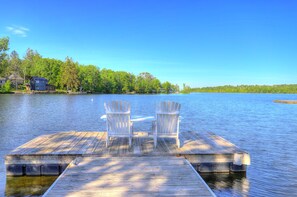 A wooden dock with two Adirondack chairs facing a calm lake, surrounded by trees and a house in the background. - Village Walk | Convenient & Charming, Hot tub (Rangeley)