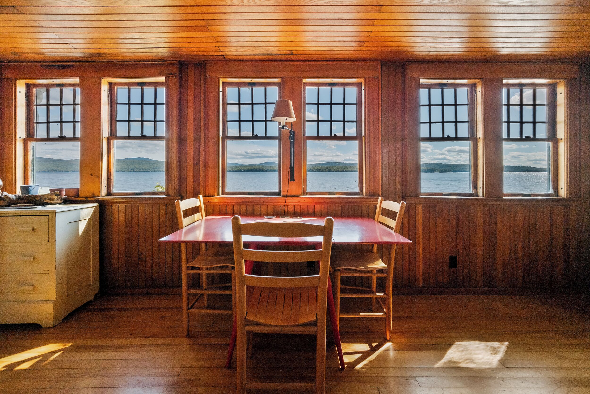 A wooden-paneled room with a dining table and chairs, large windows with a view of a lake and mountains, and a lamp on the table.