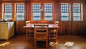 Ein Zimmer mit Holzwänden, einem Esstisch und Stühlen, großen Fenstern mit Blick auf einen See und Berge sowie einer Lampe auf dem Tisch.