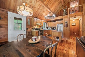 Ein Holzblockhaus-Innenraum mit Esstisch, Stühlen, einer Küchenzeile und einem Schlafbereich mit Deckenventilator.