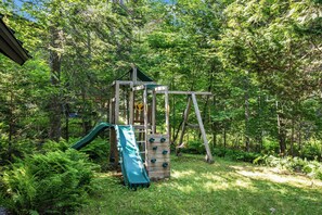 Una construcció de fusta per jugar amb tobogan i zona de paret a un pati envoltat de bosc.