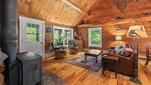 A cozy cabin interior with a wood stove, leather furniture, and large windows. - CedarDuft Cottage - Cozy log cabin right on the shores of Quimby Pond (Rangeley)