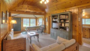 Ein gemütliches Blockhaus-Interieur mit Holzbalkendecke, einer Couch, einem Sessel, einem kleinen Tisch und einem Bücherregal.