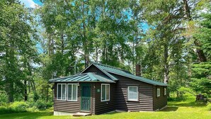 Eine kleine Holzhütte mit grüner Tür, Fenstern und geneigtem Dach, umgeben von Bäumen und Gras.