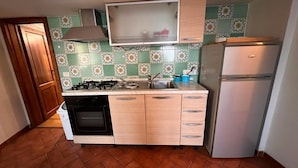 Eine kleine Küche mit Gasherd, Backofen und Kühlschrank. Der Boden ist mit einem Fischgrätmuster ausgelegt, und die Wandfliesen zeigen ein Blumenmuster.